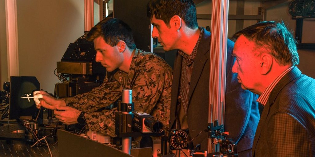 Naval Postgraduate School faculty researchers Brij Agrawal, right, and Leonardo Herrera, center, observe U.S. Navy Ensign Nicholas Messina as he positions a Reaper drone model in an optical beam path that simulates how a laser weapon system engages a distant flying drone. (Javier Chagoya)